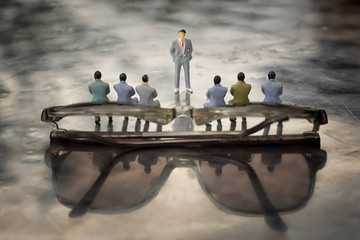 Miniature model of businessmen sitting on glasses backside and a presenter standing in front of...