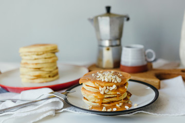 Pancakes with salted caramel and peanuts near coffee maker on white table in kitchen. Morning breakfast at home in quarantine. Recipe of homemade delicious pancakes.