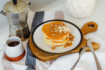 Pancakes with salted caramel and peanuts near coffee maker on white table in kitchen. Morning breakfast at home in quarantine. Recipe of homemade delicious pancakes.