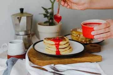 Girl pouring raspberry sauce on pancakes near coffee maker on white table in kitchen. Morning breakfast at home in quarantine. Recipe of homemade delicious pancakes.