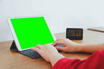 Image of hands typing on a keyboard attached to a tablet with green screen. Wood table with clock and white wall.