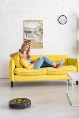 Obraz premium Mother and daughter with laptop lying on sofa near coffee table and robotic vacuum cleaner on floor in living room