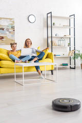 Mother smiling, hugging daughter with laptop on sofa near coffee table and looking at robotic vacuum cleaner on floor in living room