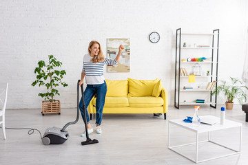 Woman with raised hand and vacuum cleaner looking away and smiling in living room