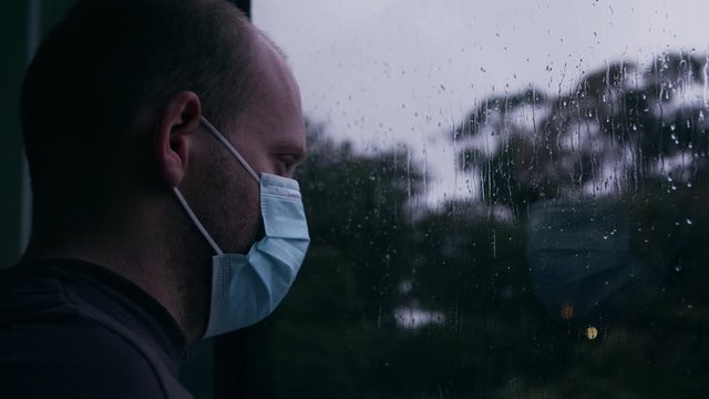 Young Man With Face Mask Looks out Window on Cold Rainy Day. Close Up 