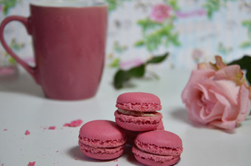 Three pink almond macaroons and mug with coffee on a white wooden background next to a pale pink rose. good morning concept