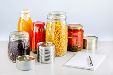 Fototapeta na wymiar Different glass jars with grains, pickled vedetables, pasta, cans of canned food. Groceries with list on the table. Food supplies crisis food stock for quarantine isolation period.