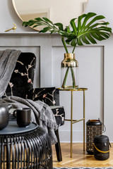Modern concept of home staging with design armchair, round mirror, black rattan coffee table, tea pot, lanterns, leaves in vase, gold table and elegant personal accessories at stylish living room.