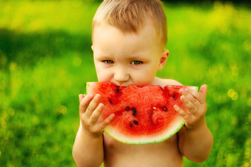 Portrait of little kid without t-shirt in the village, on the fresh air. Two years old
child eating piece of watermelon on the grass in garden. Dirty face, happy and emotional
kid, juicy watermelon.