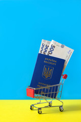 Ukrainian biometric passport with hryvnias in mini shopping cart on blue-yellow background,...