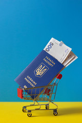 Ukrainian biometric passport with hryvnias in mini shopping cart on blue-yellow background, vertical orientation