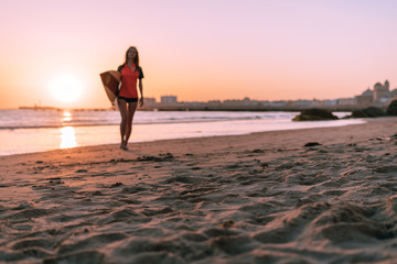 Against the backdrop of a magnificent ocean sunset, a girl is walking with a surfboard under her arm. A big city can be seen in the distance.