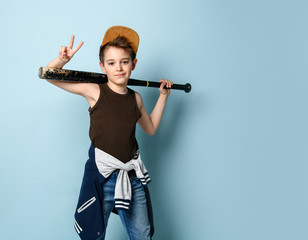 Teenager in cap and casual clothes. He holding black baseball bat, smiling and showing victory sign. Posing on blue background