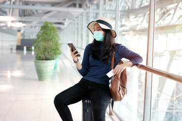 Young Asian passenger sit on black luggage with mask, using smartphone to shopping and search airline, hotel accommodation website for booking, holding credit card in hand for online reduction payment