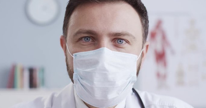 Close-up portrait of smart blue-eyed therapist with beard in white gown with statoscope and medical mask looking at camera. Male doctor in respiratory protection in cabinet. Сoncept of pandemic