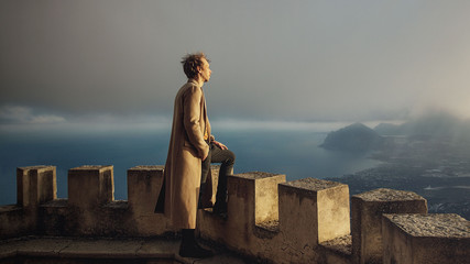 A young man in a brown coat standing on the top of the castle looking down to the a view of sea and...