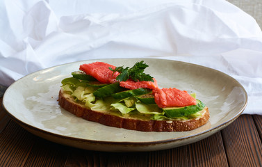 Large toast of rye bread with cheese, cabbage, parsley, fresh cucumber and red fish salmon on wooden background with white cloth drapery.