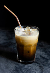 Iced coffee with milk in a tumbler glass cup with a metal straw on a dark background