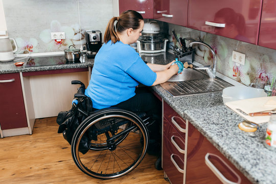 Disabled young woman in wheelchair washing dishes in specially equipped kitchen