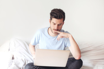 Handsome casual young man in bed with laptop. Solving business troubles in a distance. Concept of work in isolation