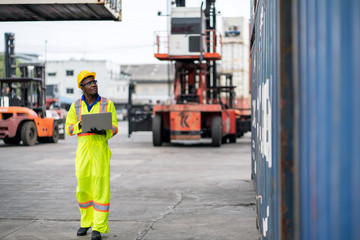 Fototapeta premium African technician dock worker in protective safety jumpsuit uniform and with hardhat and usel aptop computer at cargo container shipping warehouse. transportation import,export logistic industrial