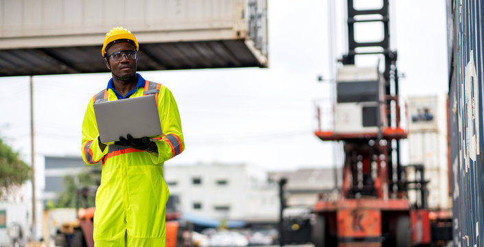 African technician dock worker in protective safety jumpsuit uniform and with hardhat and usel aptop computer at cargo container shipping warehouse. transportation import,export logistic industrial