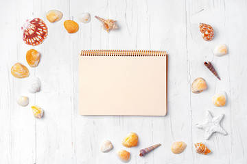 Open notepad with sea shells on white wooden background, top view
