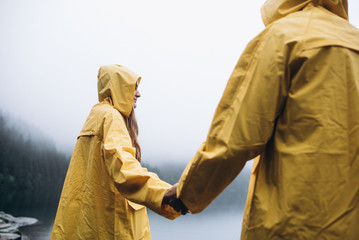 A man and a woman in yellow raincoats walk near a lake in the mountains in foggy weather.
