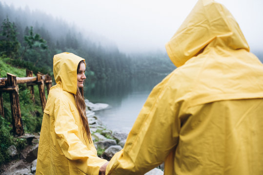 A woman in yellow raincoats holds a man's hand near a lake in the mountains in foggy weather.