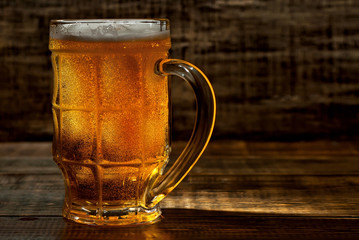 A glass of amber beer. Glasses on a wooden table. Black shabby boards on the background. Bright contrasting light and glare on the glass. Place for text near the booze.