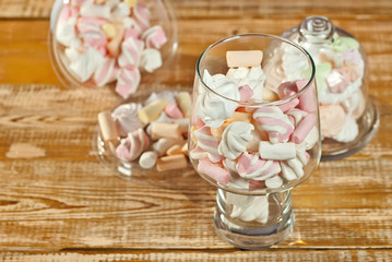 Sweets in a glass on a wooden table. Transparent utensils with marshmallows and biz on a shabby old board. Pink with white soft candies close up.