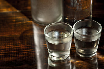 Frozen glasses with alcohol near the bottle. Drinks on a dark brown table. Transparent utensils on a black background. Old shabby boards under glass.
