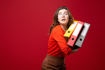 The girl in glasses carries folders, with a fright on her face on a red background. Mood stress,...