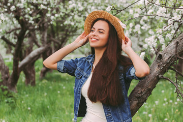 Portrait beautiful carefree hipster woman in stylish straw hat and denim jacket relax and have fun in green blossom park. Young happy hippie girl posing with long hair and smile on countryside garden