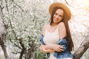 Portrait happy gentle hippie woman in stylish denim jacket and straw hat posing on countryside garden with blossom trees and smiling. Young beautiful sexy hipster girl relax in green spring sunny park