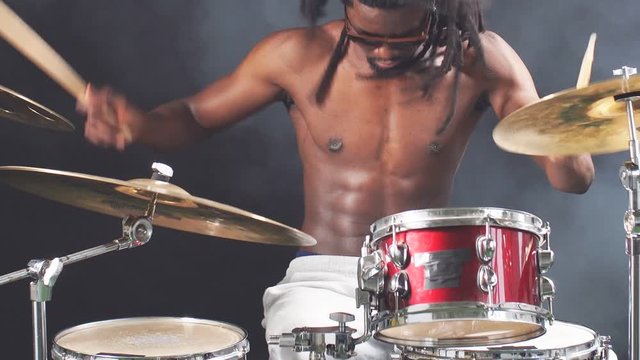 Young african man with dreadlocks sit beating on drums with sticks, with naked skin isolated over smoky background