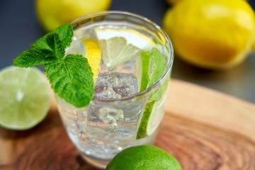 Top view image of cold refreshing summer limonade, mojito or gin tonic with fresh mint and ice cubes, lime and lemon on colorful wooden table.