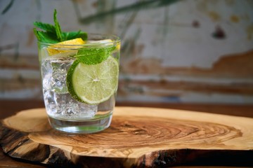 Cold refreshing summer limonade, mojito or gin tonic in glass, with fresh mint and ice cubes, lime and lemon on wooden board. Summer cocktails and drinks concepts. Copy space for text.