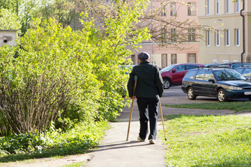 Fototapeta na wymiar A lonely elderly man with a walking stick walks through the yard of an apartment building in a park among green trees and parked cars.