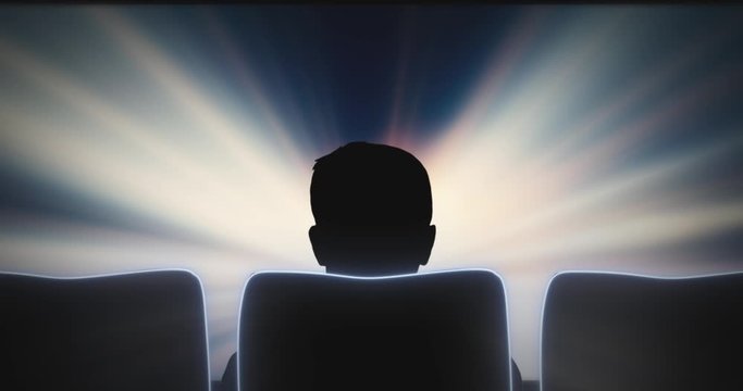 Rear view of the silhouette of a person in a cinema chair looking at a glowing screen. Zoom-in