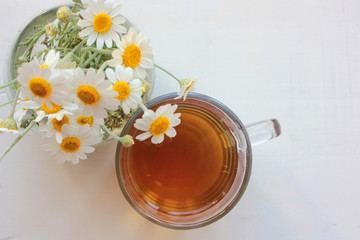 Chamomile herbal tea in glass cup and bouquet of white daisies in a glass vase on the white wooden background with copy space. Top view