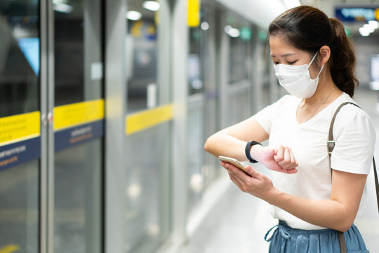 young asian woman wearing face mask, using smart phone and standing at the gate station, waiting for metor during covid-19 or coronavirus outbreak. social distancing and new normal lifestyle concept