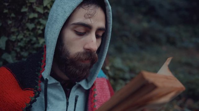 Young hipster hooded man with blanket is sitting under a tree in the woods and turning pages of an old book . Rainy cold winter day