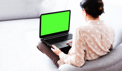 Middle-aged brunette woman on the gray sofa working on green screen laptop, soft focus, stay at home concept, cozy background.