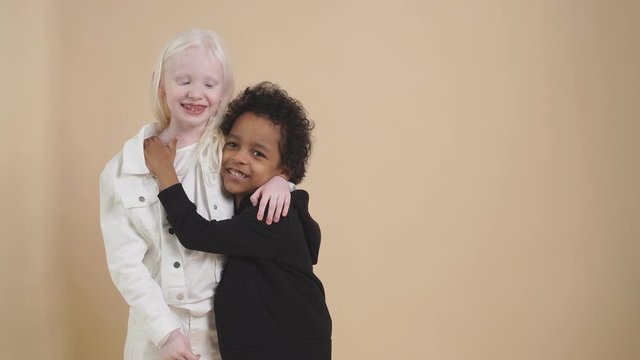 Two friendly american boy and albino children stand together isolated over beige background, diverse positive boy and girl stand hugging. Children concept.
