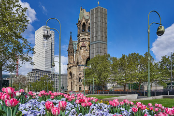 berlin germany, The Kaiser Wilhelm Memorial Church, in GermanKaiser-Wilhelm-Gedächtniskirche, spring time in the city, with a blue sky and colourful flowers, green trees on the road side - obrazy, fototapety, plakaty