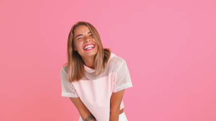 Blonde woman with shiny freckles, dressed in top. She is dancing, laughing covering her mouth with hand, posing against pink background. Close up