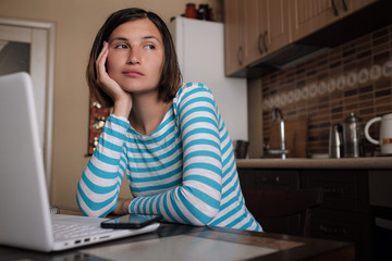 Young woman sitting in kitchen and working on laptop in morning. Female using laptop dining table