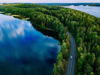 Aerial view of road with cars between green forest and blue lake water in Finland