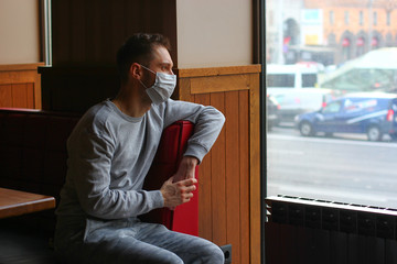The man in the blue protective medical mask is sitting at home at the window on quarantine and looking to the street